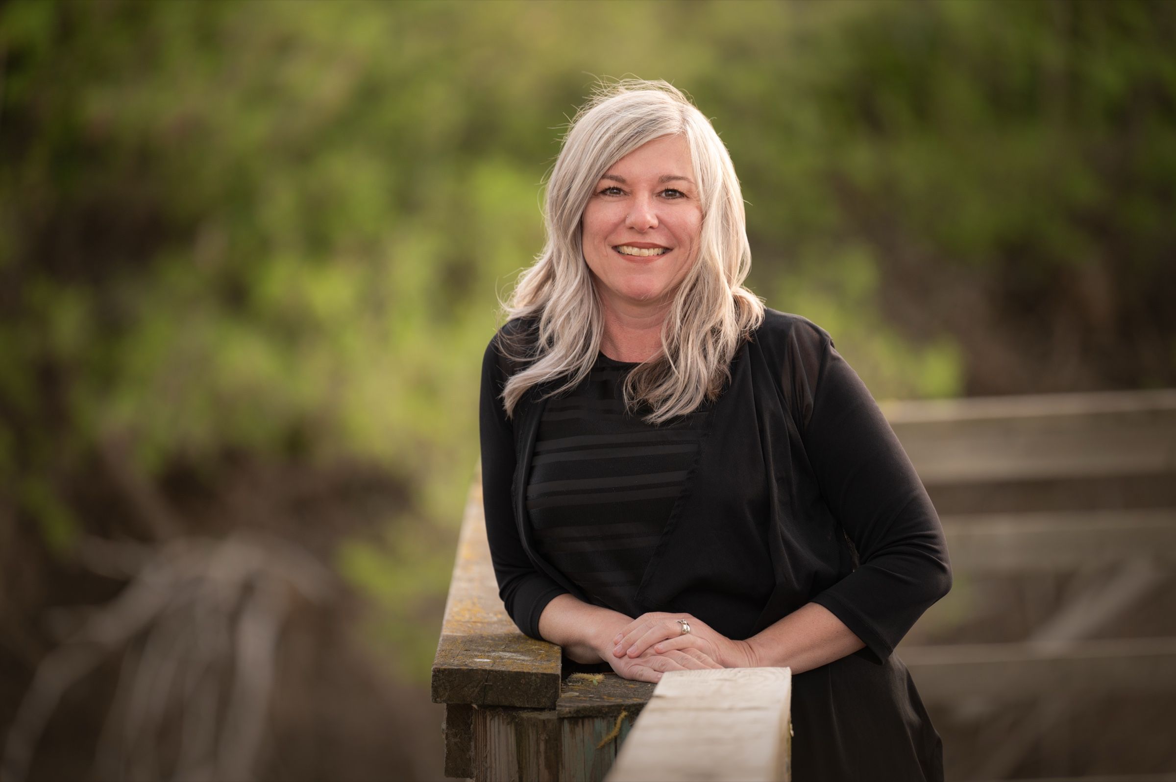 Cappi Bowers smiling, leaning on a weathered boardwalk railing with green trees behind her
